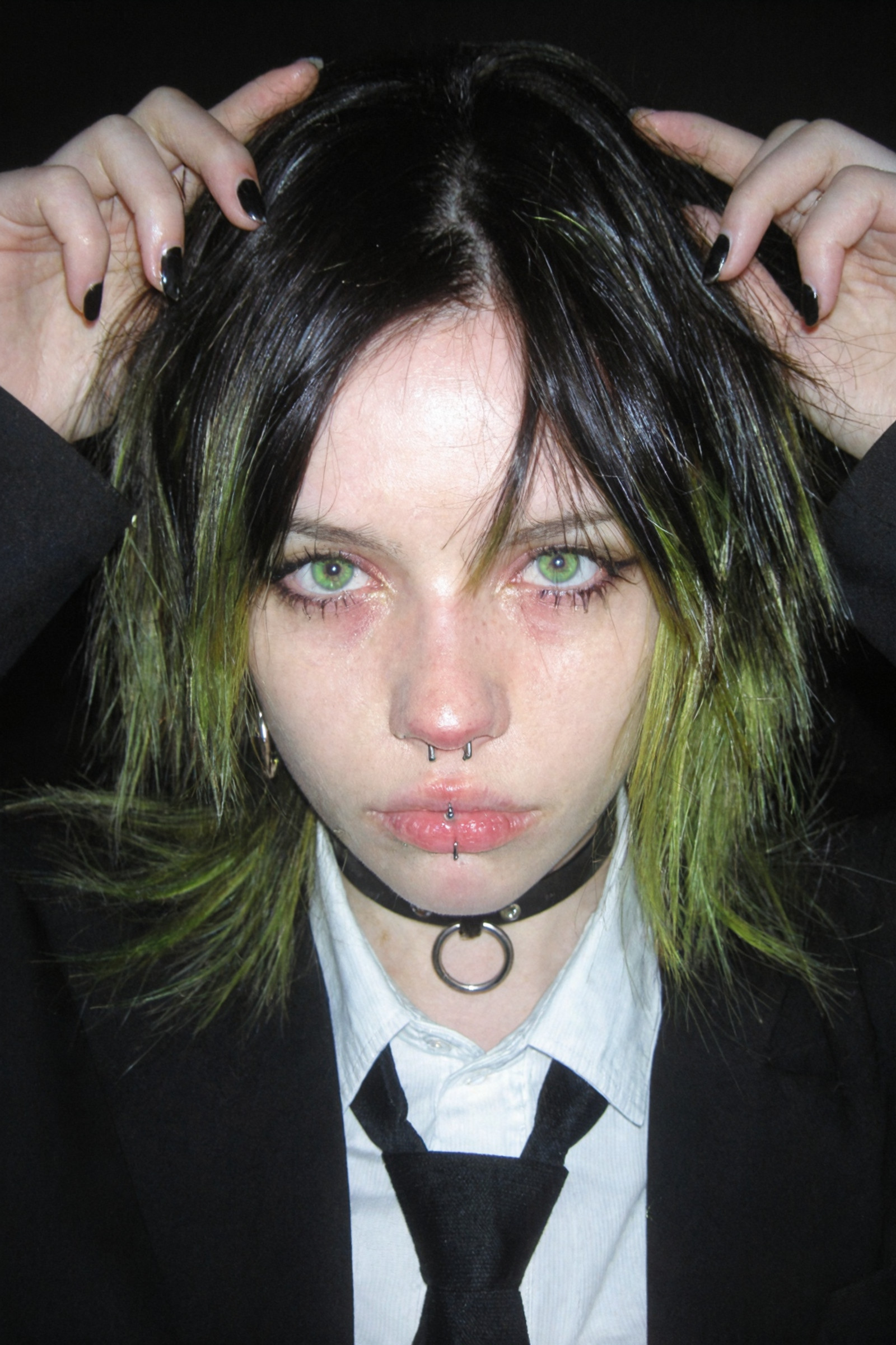 Hyper-realistic amateur candid photograph, raw 35mm flash photography style, shot on Fujifilm Superia 400. A young woman with a rebellious grunge aesthetic, messy layered wolf-cut hair transitioning from jet black roots to vibrant toxic lime green tips, hair appears slightly greasy and unkempt with flyaways caught in the harsh direct flash. She is wearing an ill-fitting oversized black suit jacket, a crisp white collared dress shirt slightly unbuttoned at the top, and a loose black necktie.

Extreme close-up macro detail on facial features: pale, translucent skin with visible subsurface scattering, heavy natural blush and rosacea on the cheeks and nose bridge giving a feverish appearance, distinct skin pores, uneven skin texture, sweat sheen on the upper lip, and fine vellus peach fuzz on the jawline. Piercing neon green eyes with dilated pupils and visible red capillaries in the sclera, heavy hooded eyelids with dark under-eye circles indicating exhaustion. A silver circular barbell lip piercing (snake bite or center labret) indenting the soft tissue of the lower lip, silver hoop earrings, and a tight black leather choker with a silver O-ring compressing the neck skin.

She is posing with both hands raised to head height, palms facing the camera, fingers slightly curled in a casual gesture. Fingernails are painted with chipped, glossy black polish, revealing the nail bed underneath, visible cuticles and dry skin around the fingertips. The lighting is a harsh on-camera flash creating a stark contrast against a pitch-black void background, hard rim lighting, slight chromatic aberration, film grain, vignette, 8k resolution, photorealistic textures, uncompromising realism.