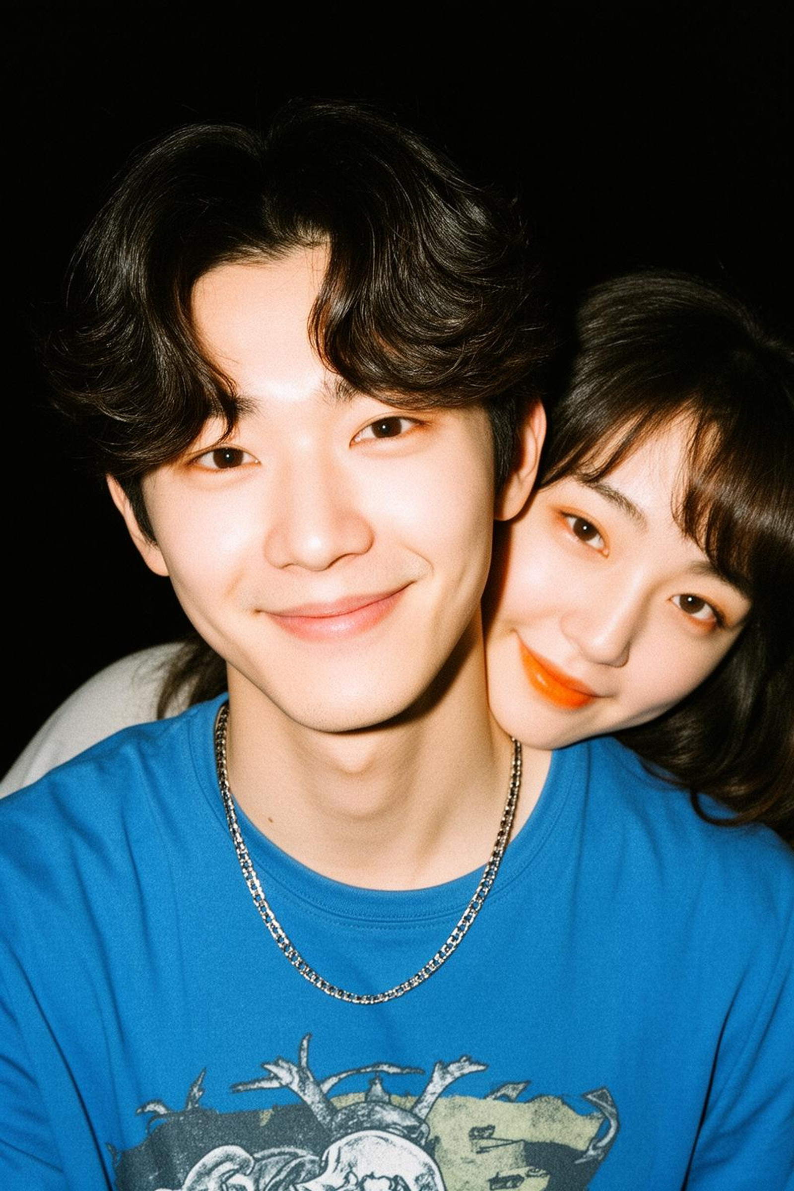 Close-up photograph capturing an intimate moment between a young Korean couple. The man is in the foreground. He has wavy, center-parted dark hair and a gentle smile, wearing a blue graphic t-shirt and a silver chain necklace. A woman leans in from behind, resting her chin on his left shoulder, her face framed by soft dark hair as she looks warmly toward the camera. Both have light makeup with soft orange-tinted lips. The lighting is reminiscent of a candid flash-photography style, creating high contrast with a dark, shadowy background and slightly grainy texture. The composition is tight and centered, focusing on their facial expressions and the physical closeness between them. The overall mood is affectionate, casual, and nostalgic, like a personal social media snapshot.<lora:TA-2026-02-27-22-04-54-Close-upph-82:0>