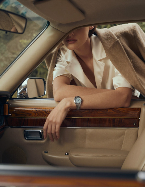 A high-fashion editorial advertising shot inside a vintage American luxury car with a rich retro atmosphere. The perspective is from the driver’s point of view, looking toward the passenger seat. The frameless car door acts like a natural cinematic frame, precisely enclosing the composition.

The interior of the car is rendered in ultra-realistic detail: smooth premium leather seats with subtle creases and natural wear, polished wood paneling with deep warm tones and visible grain, brushed metal accents reflecting soft highlights. The dashboard and door trim carry a muted vintage palette — caramel brown, dark walnut, and soft champagne metallics — all under controlled, diffused lighting.

Outside the car, a woman leans forward against the open window. Her posture feels effortless and intentional — one arm laid horizontally along the lower edge of the window frame, fingers relaxed with natural tension, while her other arm bends upward, gently supporting her chin. The pose creates elegant lines and a sense of quiet intimacy with the viewer.

The composition deliberately crops her face, revealing only the lower half — soft lips, smooth skin, and a refined jawline, emphasizing mystery and allure. Her styling is impeccable: she wears tailored, high-end garments with precise structure and luxurious fabrics — fine wool, silk blends, or structured satin, showing realistic folds, tension, and light absorption. The stitching, seams, and edges are crisp and believable.

On her wrist, an expensive women’s watch catches the light — polished metal, sapphire glass reflections, and a finely detailed dial visible at close range, reinforcing the commercial luxury theme.

Lighting is soft yet directional, like controlled studio light blended with natural daylight, producing gentle highlights on skin, fabric, and car surfaces. Subtle reflections on the window edge and chrome details enhance realism. The color palette is restrained and sophisticated: warm browns, champagne gold, soft ivory, and muted skin tones, with slight film-like desaturation for a vintage editorial feel.

Style: ultra-realistic, high-fashion photography, commercial luxury advertising, cinematic framing, shallow depth of field (focus on lips, hand, and watch), 85mm lens look, soft bokeh, fine film grain, high dynamic range, extremely detailed textures — leather grain, fabric weave, skin pores, metallic reflections. Mood: elegant, intimate, restrained, quietly seductive, timeless.