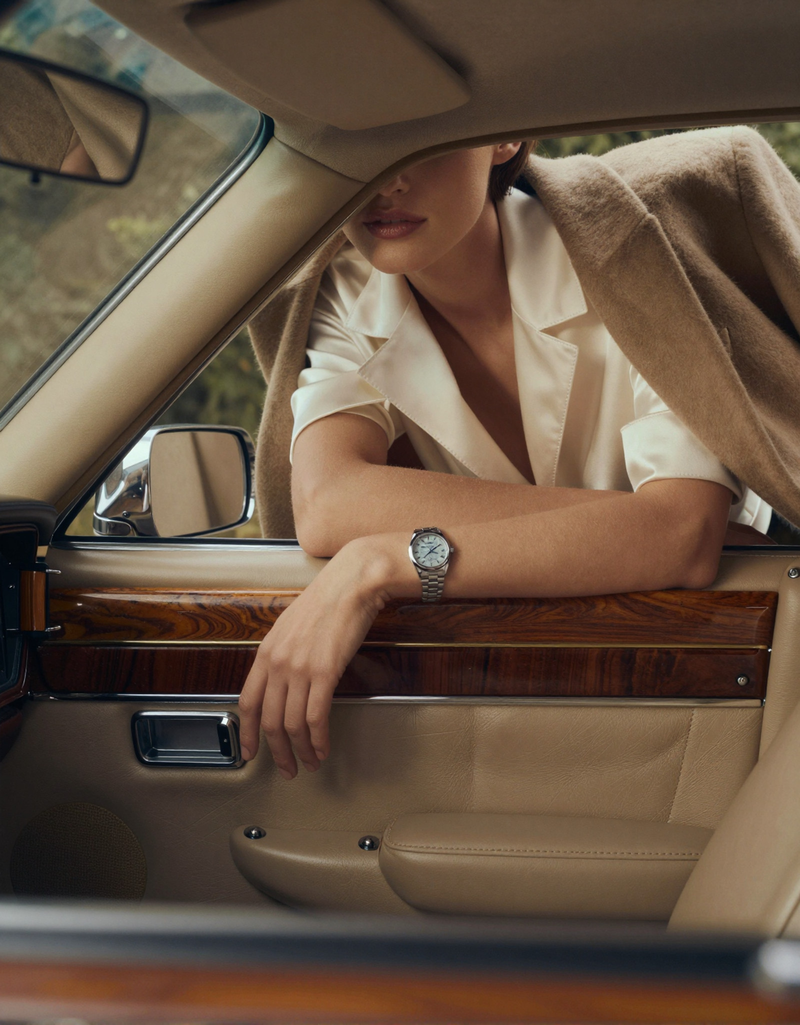 A high-fashion editorial advertising shot inside a vintage American luxury car with a rich retro atmosphere. The perspective is from the driver’s point of view, looking toward the passenger seat. The frameless car door acts like a natural cinematic frame, precisely enclosing the composition.

The interior of the car is rendered in ultra-realistic detail: smooth premium leather seats with subtle creases and natural wear, polished wood paneling with deep warm tones and visible grain, brushed metal accents reflecting soft highlights. The dashboard and door trim carry a muted vintage palette — caramel brown, dark walnut, and soft champagne metallics — all under controlled, diffused lighting.

Outside the car, a woman leans forward against the open window. Her posture feels effortless and intentional — one arm laid horizontally along the lower edge of the window frame, fingers relaxed with natural tension, while her other arm bends upward, gently supporting her chin. The pose creates elegant lines and a sense of quiet intimacy with the viewer.

The composition deliberately crops her face, revealing only the lower half — soft lips, smooth skin, and a refined jawline, emphasizing mystery and allure. Her styling is impeccable: she wears tailored, high-end garments with precise structure and luxurious fabrics — fine wool, silk blends, or structured satin, showing realistic folds, tension, and light absorption. The stitching, seams, and edges are crisp and believable.

On her wrist, an expensive women’s watch catches the light — polished metal, sapphire glass reflections, and a finely detailed dial visible at close range, reinforcing the commercial luxury theme.

Lighting is soft yet directional, like controlled studio light blended with natural daylight, producing gentle highlights on skin, fabric, and car surfaces. Subtle reflections on the window edge and chrome details enhance realism. The color palette is restrained and sophisticated: warm browns, champagne gold, soft ivory, and muted skin tones, with slight film-like desaturation for a vintage editorial feel.

Style: ultra-realistic, high-fashion photography, commercial luxury advertising, cinematic framing, shallow depth of field (focus on lips, hand, and watch), 85mm lens look, soft bokeh, fine film grain, high dynamic range, extremely detailed textures — leather grain, fabric weave, skin pores, metallic reflections. Mood: elegant, intimate, restrained, quietly seductive, timeless.