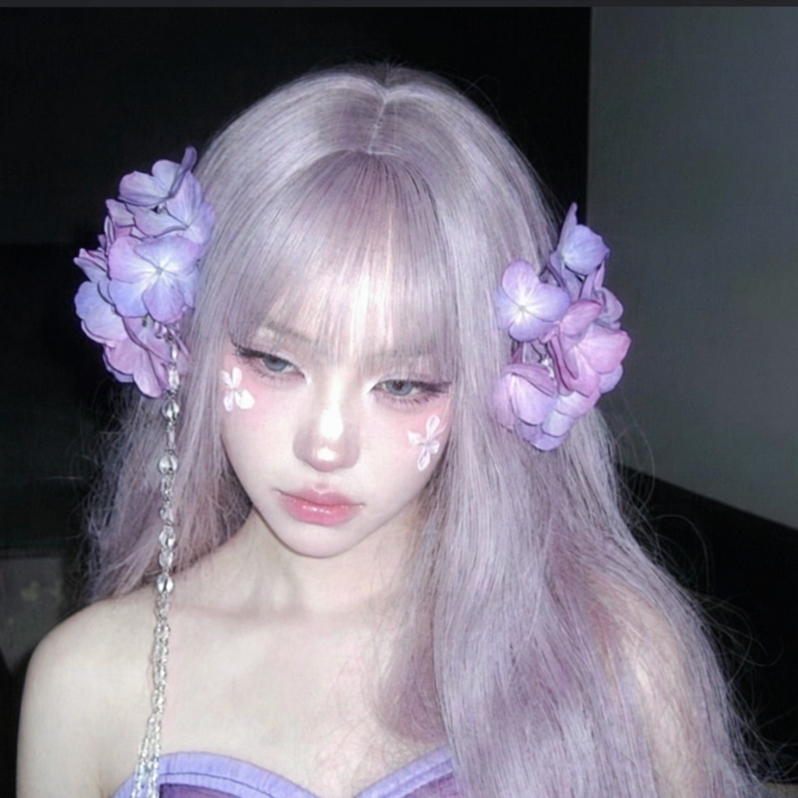 Portrait of a young woman, pale porcelain skin, voluminous light lavender hair, thick bangs, synthetic wig texture. Stylized makeup, heavy pink under-eye blush (igari style), painted white floral petal decals on cheeks, sharp downward eyeliner, glossy pale pink lips, pale blue contact lenses, melancholic gaze. Large artificial purple hydrangea hair accessories, dangling clear crystal bead strings. Strapless lavender garment. Harsh direct camera flash, hard shadows, pitch black background, amateur candid aesthetic, raw 35mm film grain, high contrast, detailed skin pores, unpolished realism.