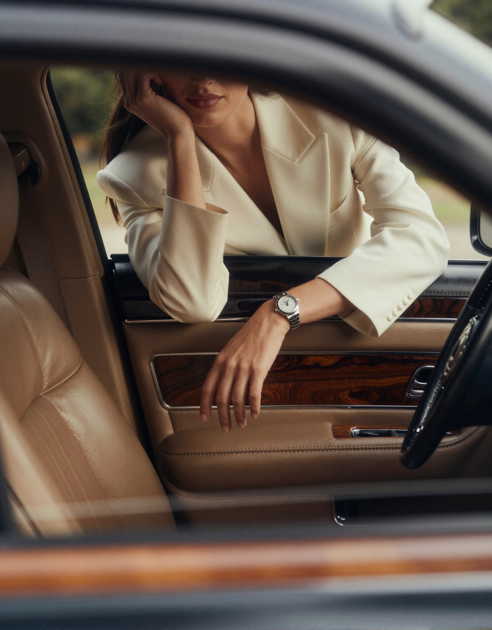 A high-fashion editorial advertising shot inside a vintage American luxury car with a rich retro atmosphere. The perspective is from the driver’s point of view, looking toward the passenger seat. The frameless car door acts like a natural cinematic frame, precisely enclosing the composition.

The interior of the car is rendered in ultra-realistic detail: smooth premium leather seats with subtle creases and natural wear, polished wood paneling with deep warm tones and visible grain, brushed metal accents reflecting soft highlights. The dashboard and door trim carry a muted vintage palette — caramel brown, dark walnut, and soft champagne metallics — all under controlled, diffused lighting.

Outside the car, a woman leans forward against the open window. Her posture feels effortless and intentional — one arm laid horizontally along the lower edge of the window frame, fingers relaxed with natural tension, while her other arm bends upward, gently supporting her chin. The pose creates elegant lines and a sense of quiet intimacy with the viewer.

The composition deliberately crops her face, revealing only the lower half — soft lips, smooth skin, and a refined jawline, emphasizing mystery and allure. Her styling is impeccable: she wears tailored, high-end garments with precise structure and luxurious fabrics — fine wool, silk blends, or structured satin, showing realistic folds, tension, and light absorption. The stitching, seams, and edges are crisp and believable.

On her wrist, an expensive women’s watch catches the light — polished metal, sapphire glass reflections, and a finely detailed dial visible at close range, reinforcing the commercial luxury theme.

Lighting is soft yet directional, like controlled studio light blended with natural daylight, producing gentle highlights on skin, fabric, and car surfaces. Subtle reflections on the window edge and chrome details enhance realism. The color palette is restrained and sophisticated: warm browns, champagne gold, soft ivory, and muted skin tones, with slight film-like desaturation for a vintage editorial feel.

Style: ultra-realistic, high-fashion photography, commercial luxury advertising, cinematic framing, shallow depth of field (focus on lips, hand, and watch), 85mm lens look, soft bokeh, fine film grain, high dynamic range, extremely detailed textures — leather grain, fabric weave, skin pores, metallic reflections. Mood: elegant, intimate, restrained, quietly seductive, timeless.