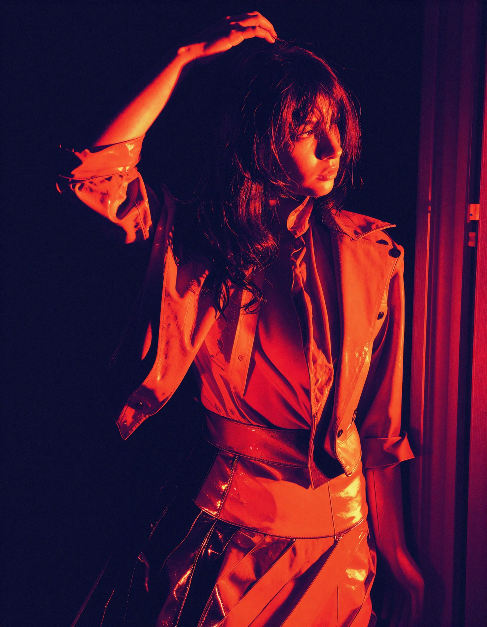 Bold sensual fashion graphic, medium portrait composition, dramatic minimalism, sharp silhouette, glossy wet shine, and a red-orange with deep plum palette.

A feminine figure stands against a dark background with her body turned slightly to the side. Scratching the top of her head. Her face is partly hidden in shadow, and her pose feels poised and self-possessed. She wears an open cropped jacket and plated skirt.

She stands on a doorway, partly lit.

Intense red-orange light washes across the raised arm, face, torso, and glossy clothing, creating a heated reflective glow. Deep plum and near-black shadows surround the figure and obscure most of the background. The image feels sleek, polished, intimate, and electrically charged.