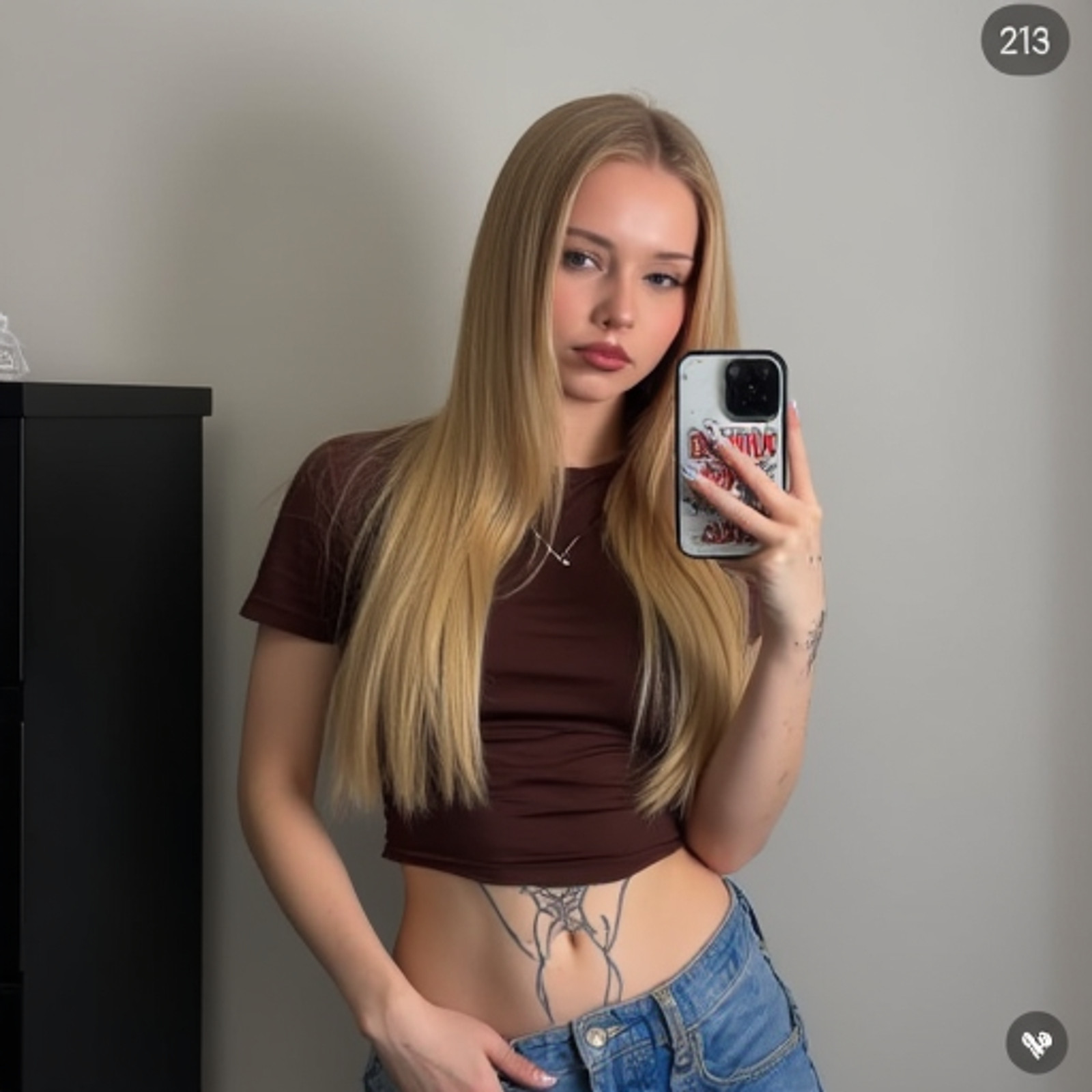 Photograph of a young Caucasian woman with long, straight blonde hair taking a mirror selfie. She has light skin, blue eyes, and is wearing a brown, short-sleeved, cropped t-shirt that reveals her midriff, showing a tattoo on her left side. She also wears blue, slightly unbuttoned jeans. Her right hand holds a smartphone with a "Rambo" movie poster case, positioned near her face. She has a subtle, pouty expression with slightly parted pink lips. Her left arm, partially visible, has another tattoo. The background is a plain, light-gray wall with a black, tall, rectangular cabinet on the left. The image has a watermark in the upper right corner with the number "213." The overall style is casual and modern, with a focus on the woman's reflection and her tattoos.