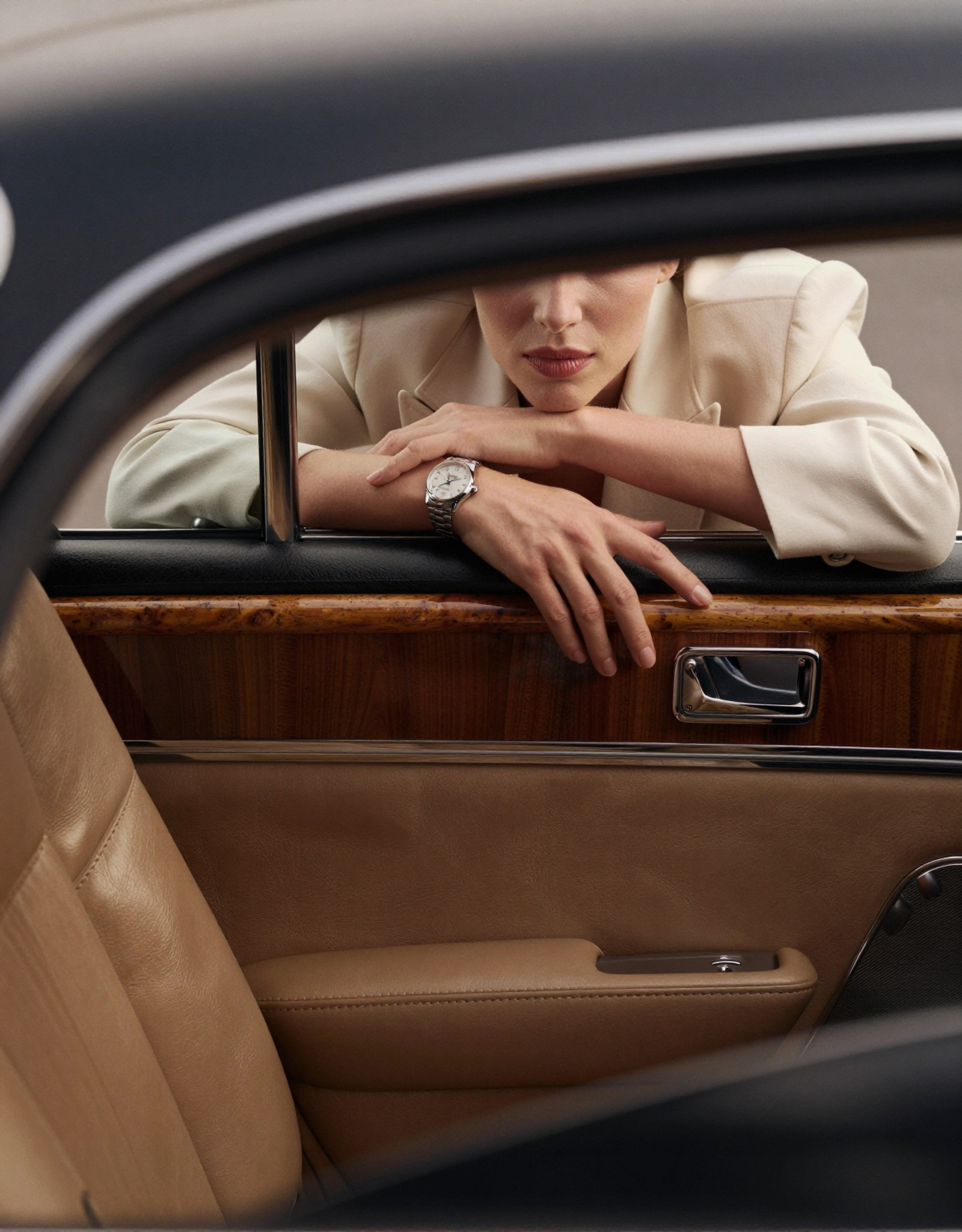 A high-fashion editorial advertising shot inside a vintage American luxury car with a rich retro atmosphere. The perspective is from the driver’s point of view, looking toward the passenger seat. The frameless car door acts like a natural cinematic frame, precisely enclosing the composition.

The interior of the car is rendered in ultra-realistic detail: smooth premium leather seats with subtle creases and natural wear, polished wood paneling with deep warm tones and visible grain, brushed metal accents reflecting soft highlights. The dashboard and door trim carry a muted vintage palette — caramel brown, dark walnut, and soft champagne metallics — all under controlled, diffused lighting.

Outside the car, a woman leans forward against the open window. Her posture feels effortless and intentional — one arm laid horizontally along the lower edge of the window frame, fingers relaxed with natural tension, while her other arm bends upward, gently supporting her chin. The pose creates elegant lines and a sense of quiet intimacy with the viewer.

The composition deliberately crops her face, revealing only the lower half — soft lips, smooth skin, and a refined jawline, emphasizing mystery and allure. Her styling is impeccable: she wears tailored, high-end garments with precise structure and luxurious fabrics — fine wool, silk blends, or structured satin, showing realistic folds, tension, and light absorption. The stitching, seams, and edges are crisp and believable.

On her wrist, an expensive women’s watch catches the light — polished metal, sapphire glass reflections, and a finely detailed dial visible at close range, reinforcing the commercial luxury theme.

Lighting is soft yet directional, like controlled studio light blended with natural daylight, producing gentle highlights on skin, fabric, and car surfaces. Subtle reflections on the window edge and chrome details enhance realism. The color palette is restrained and sophisticated: warm browns, champagne gold, soft ivory, and muted skin tones, with slight film-like desaturation for a vintage editorial feel.

Style: ultra-realistic, high-fashion photography, commercial luxury advertising, cinematic framing, shallow depth of field (focus on lips, hand, and watch), 85mm lens look, soft bokeh, fine film grain, high dynamic range, extremely detailed textures — leather grain, fabric weave, skin pores, metallic reflections. Mood: elegant, intimate, restrained, quietly seductive, timeless.