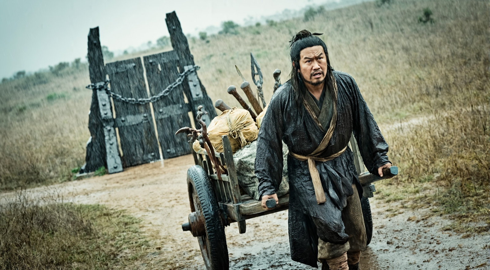 oblique view a puzzled lean mature Taiwanese male druid, pulling a wooden handcart piled high with packages and worn weapons, on a dirt track in an arid savannah landscape, in the background is mysterious trapdoor made of iron-banded timber, chained shut. heavy rain