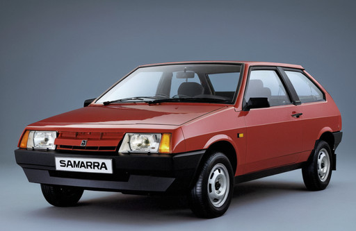 The image is a photograph of a red vaz 2108, specifically a Samara, which is a hatchback model. The car is shown from the front three-quarter angle, with the front facing towards the left side of the image. It has a sleek and modern design with a curved roofline and a sloping roofline. The front of the car has a large grille with the word "Samara" written in white letters, and the headlights are angular and angular. The wheels are black with silver rims. The background is a plain grey color, making the car stand out.<lora:Lada (VAZ)_2108_lora_XL:1>