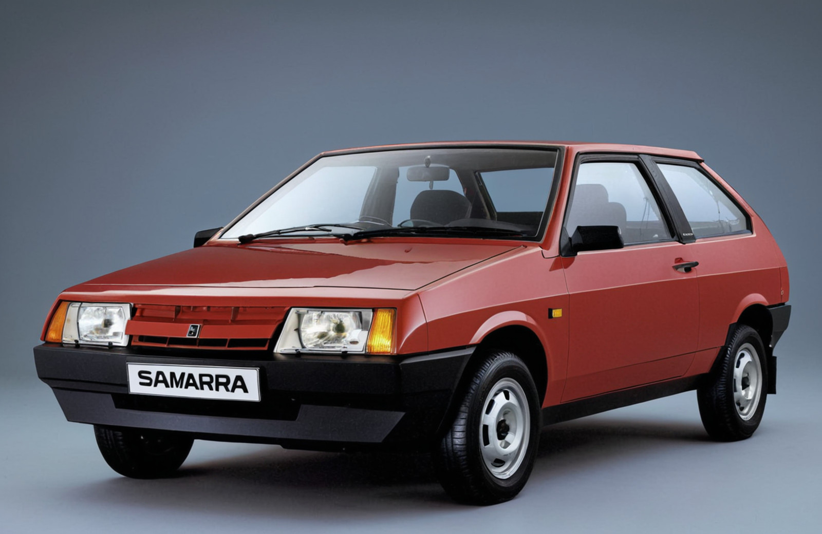 The image is a photograph of a red vaz 2108, specifically a Samara, which is a hatchback model. The car is shown from the front three-quarter angle, with the front facing towards the left side of the image. It has a sleek and modern design with a curved roofline and a sloping roofline. The front of the car has a large grille with the word "Samara" written in white letters, and the headlights are angular and angular. The wheels are black with silver rims. The background is a plain grey color, making the car stand out.<lora:Lada (VAZ)_2108_lora_XL:1>
