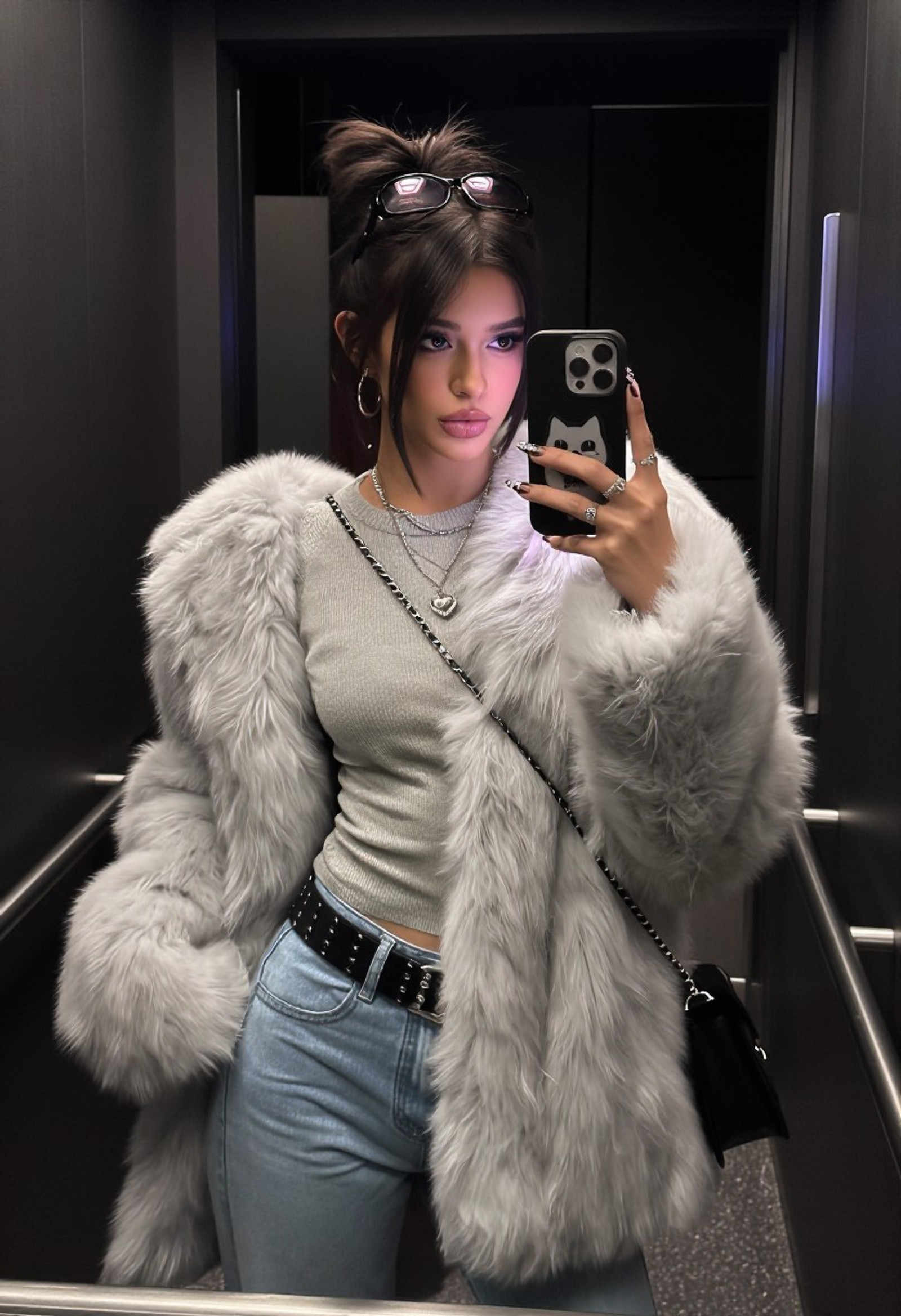 detailed texture, 8k, igmodel, hyper realistic, 
She s a young, striking woman with long, dark hair styled in a messy high bun (with face-framing strands), fair skin, sharp makeup (smoky eyes, bold brows, dark lips), and a confident, slightly sultry pout. Sunglasses are perched on her head like a headband.
She s layered in cozy winter chic: an oversized, fluffy light-gray faux fur coat (open and dramatic), over a fitted gray knit crewneck sweater, paired with high-waisted light-blue jeans cinched by a wide black studded belt. Accessories include layered necklaces (with a heart pendant), hoop earrings, multiple rings, and a black chain-strap bag slung across her body.
She s taking a full-body mirror selfie in a sleek, modern elevator or hallway (dark walls, metallic railings), holding up an iPhone with a cat-logo case. Overall vibe: luxurious, urban influencer winter fashion   plush, edgy, and effortlessly glamorous.