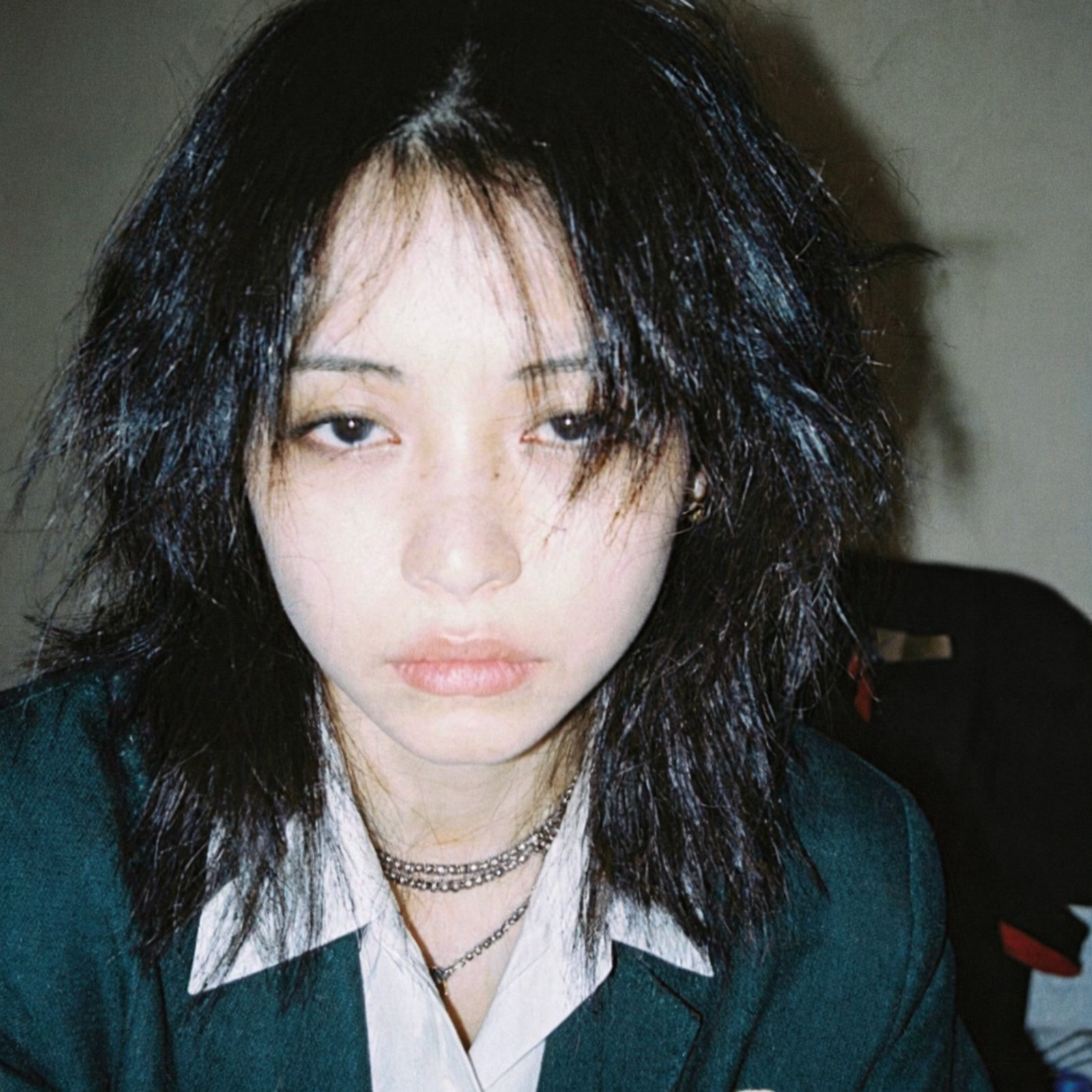 Raw amateur candid flash photography, shot on 35mm Fujifilm Superia 400, grainy low-light indoor aesthetic. A hyper-realistic interpretation of a young woman with a messy, chaotic jet-black wolf cut hairstyle, hair strands clumping together with a slightly greasy unwashed texture, wild flyaways framing the face. She has an extremely pale, translucent complexion with visible blue veins beneath the skin near the temples.

Extreme focus on facial micro-details: pronounced physiological dark circles and deep bags under the eyes (periorbital hyperpigmentation) indicating severe insomnia, heavy hooded eyelids, deadpan exhausted expression, bored sanpaku glare, dark almost black irises with sharp reflections of the camera flash. Skin texture is raw and unfiltered: visible open pores on the nose and cheeks, distinct sebaceous filaments, slight uneven skin tone, hint of sweat sheen on the forehead, peach fuzz on the jawline. Lips are pale pink, slightly chapped with vertical cracks, mouth small and set in a flat line.

She is wearing a dark teal green school uniform jacket with a white collared shirt underneath, unbuttoned at the top. Neck adorned with a layered silver beaded choker necklace and a thin metallic chain, realistic metal texture with scratches and tarnishing. Harsh direct camera flash lighting creates a hard shadow behind her against a blurred, clutter-filled bedroom background. 8k resolution, photorealistic, unedited aesthetic, harsh shadows, chromatic aberration at edges.